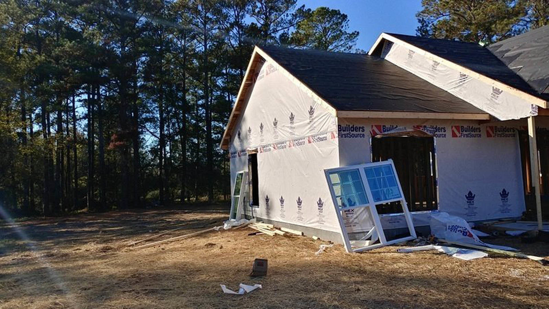 Partially built house with exposed framing, broken window leaning against exterior wall, scattered wood planks, white tent, and construction materials on dirt ground