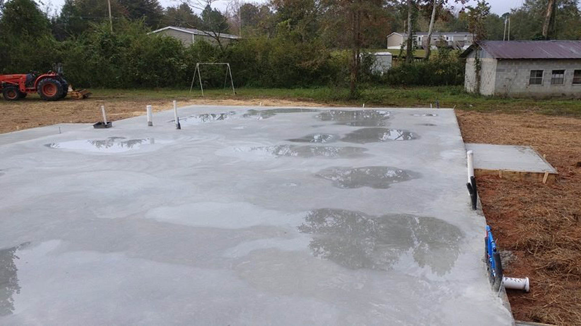 Wet concrete slab with a circular hole in the center, surrounded by grassy yard and trees, tractor partially visible in background
