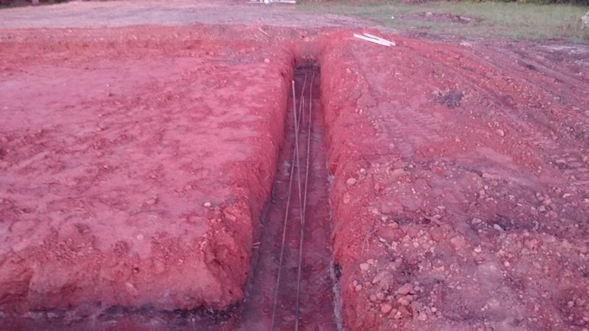 Red dirt trench with wire mesh, tire tracks, and white marking line on outdoor construction site.