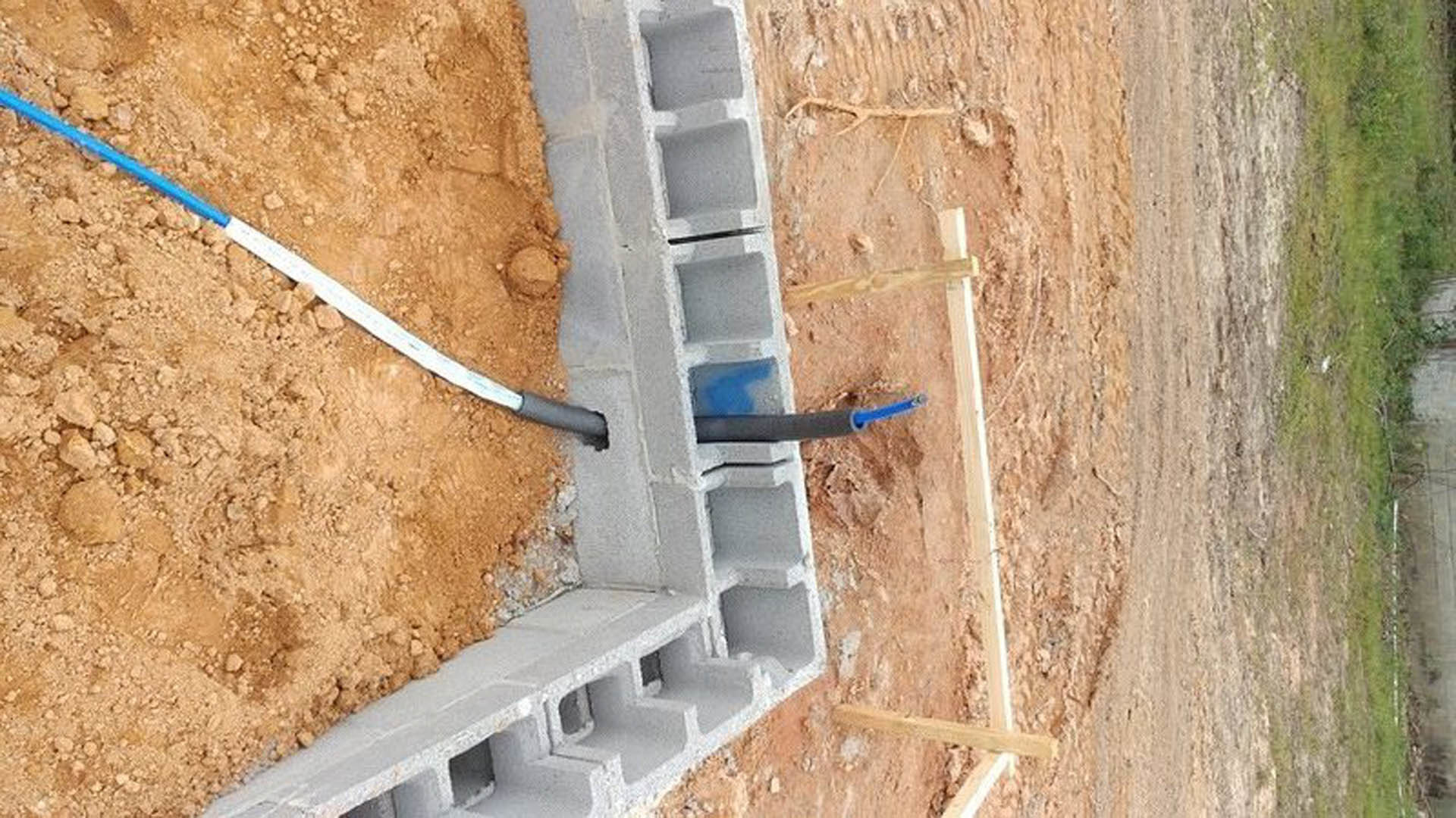 Concrete block foundation with exposed pipe, blue and white plastic stick on soil, close-up of unfinished concrete wall and dirt ground