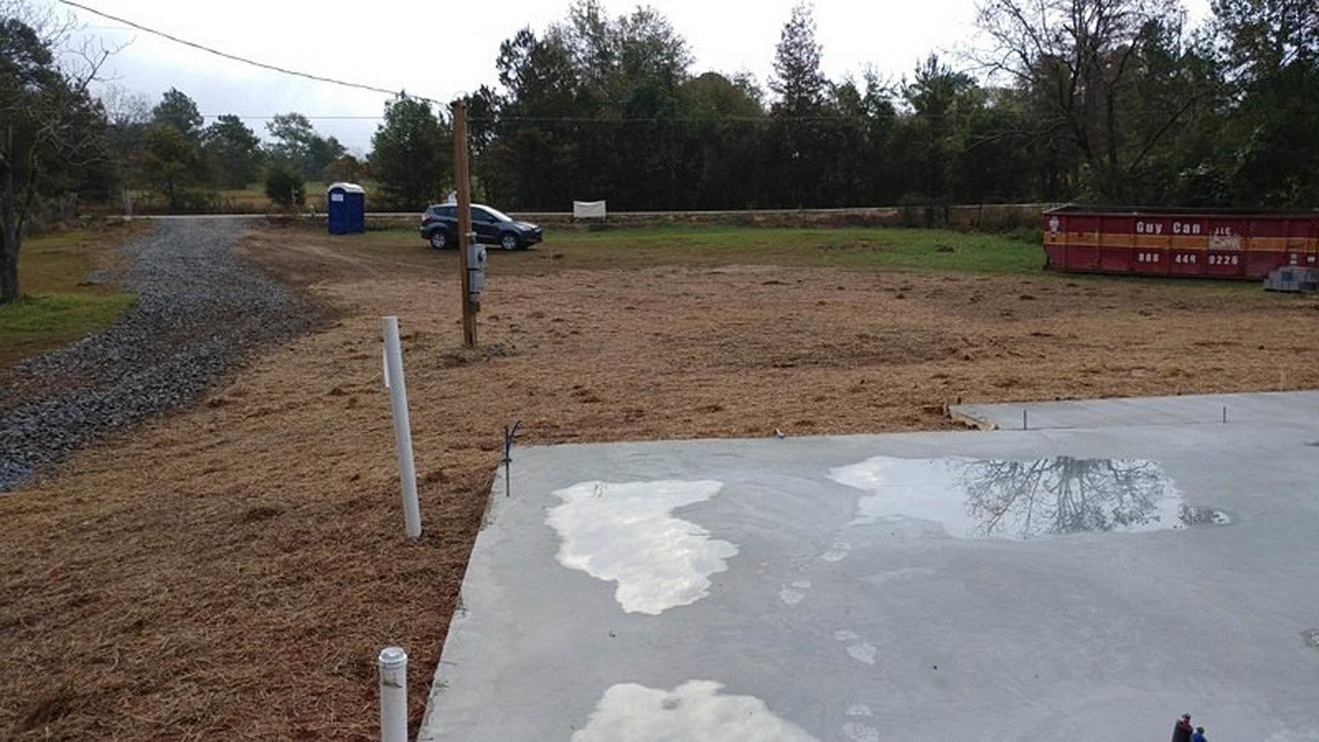 Wet concrete patio with a large puddle of water, red and yellow container with white text, blue portable toilet on grassy area, trees and sky in background