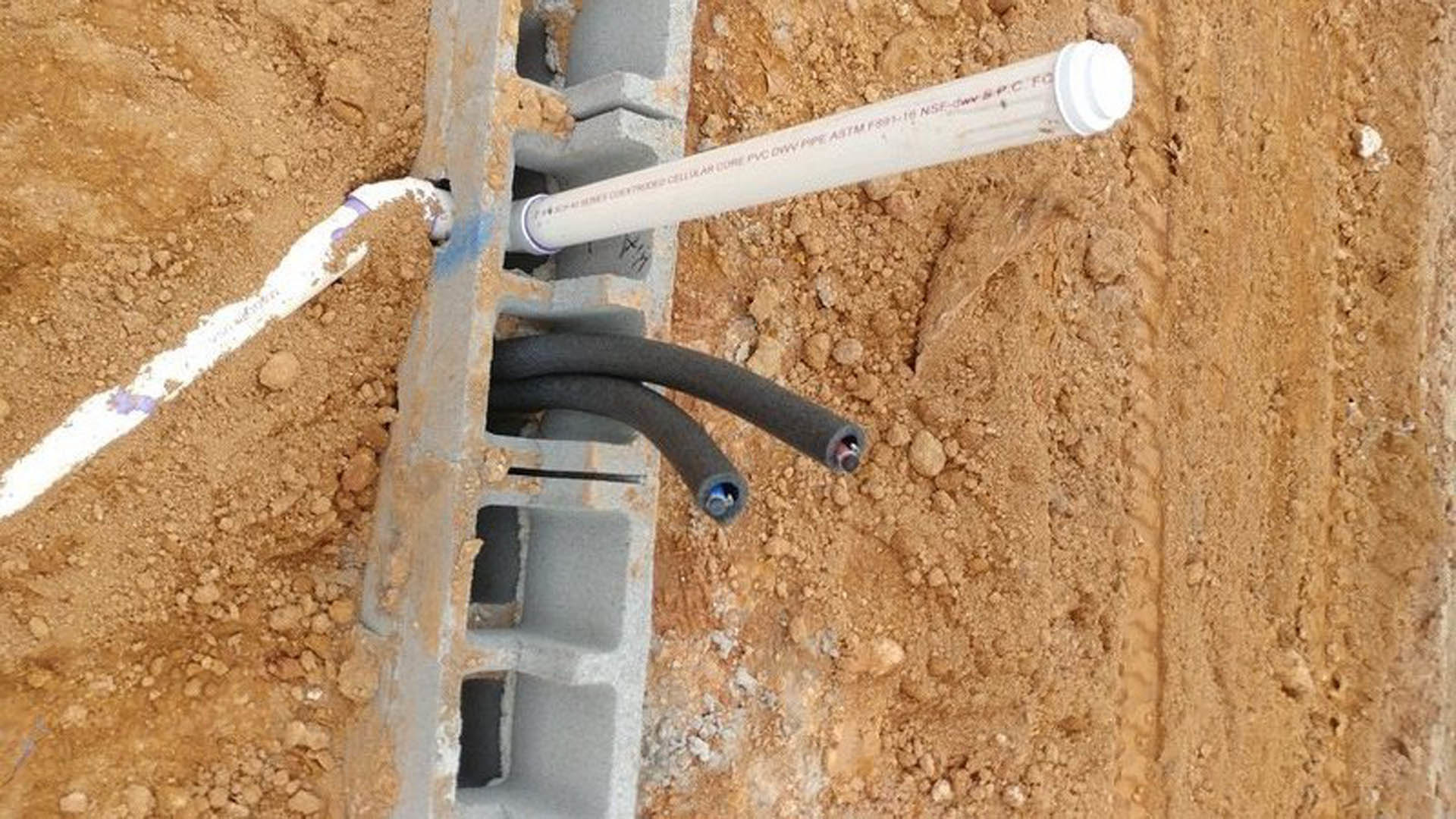 White PVC pipe emerging from dirt in a hole, surrounded by concrete block and black electrical wires outdoors