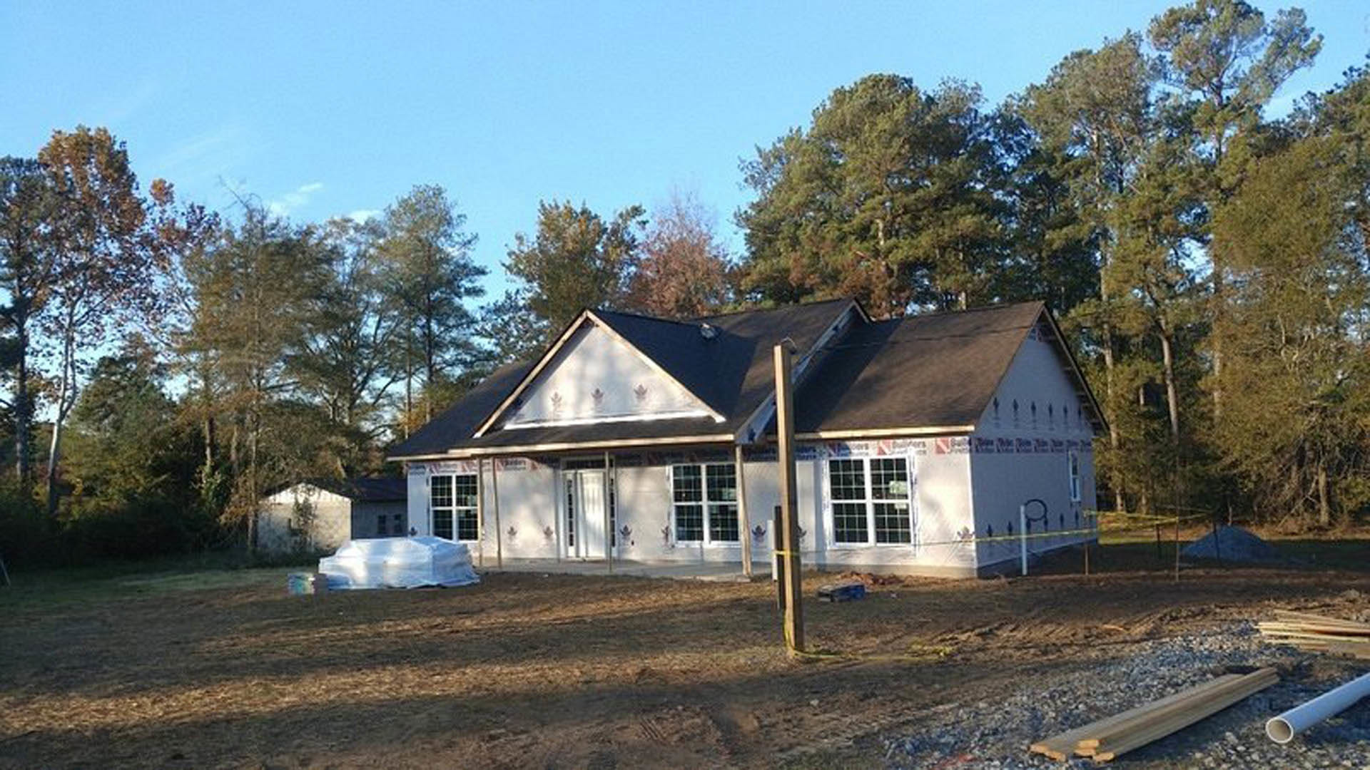 Framed house under construction with exposed plywood walls, shingled roof, and surrounding mature trees