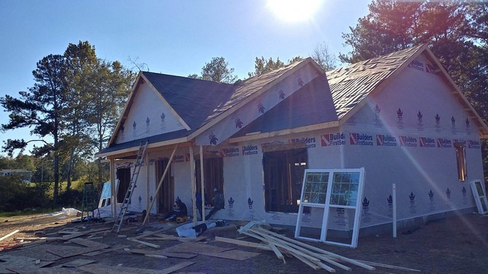 Partially built house exterior with exposed framing, white-framed window, ladder leaning against wall, tree visible in background