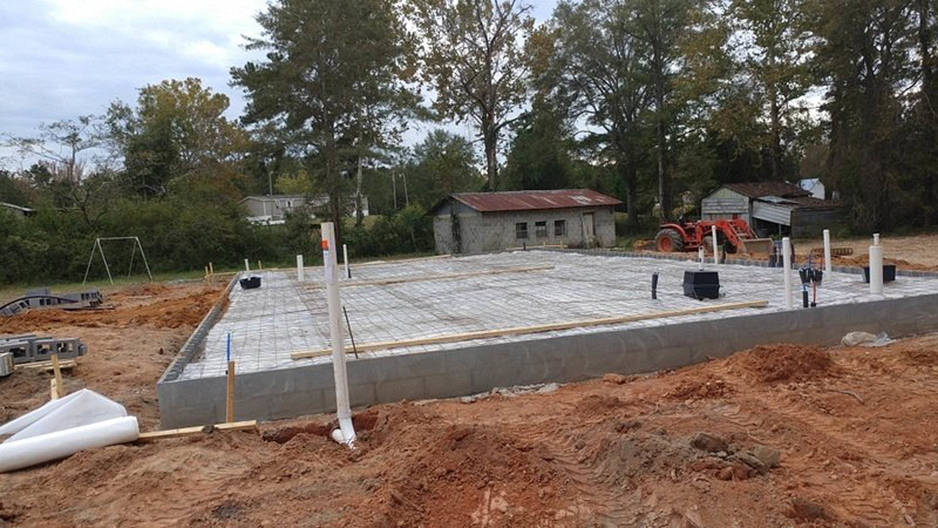 Concrete foundation and metal frame structure with red roof, red tractor parked in front, soil and trees surrounding construction site