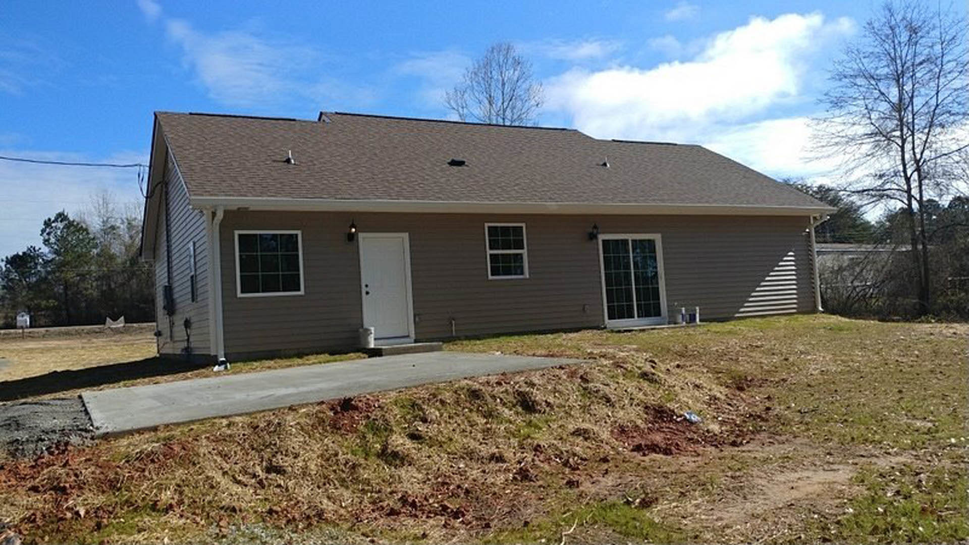 Two-story house with white framed windows, black hardware on white front door, paved driveway bordered by grass and dirt, mature trees in background, partly cloudy sky overhead