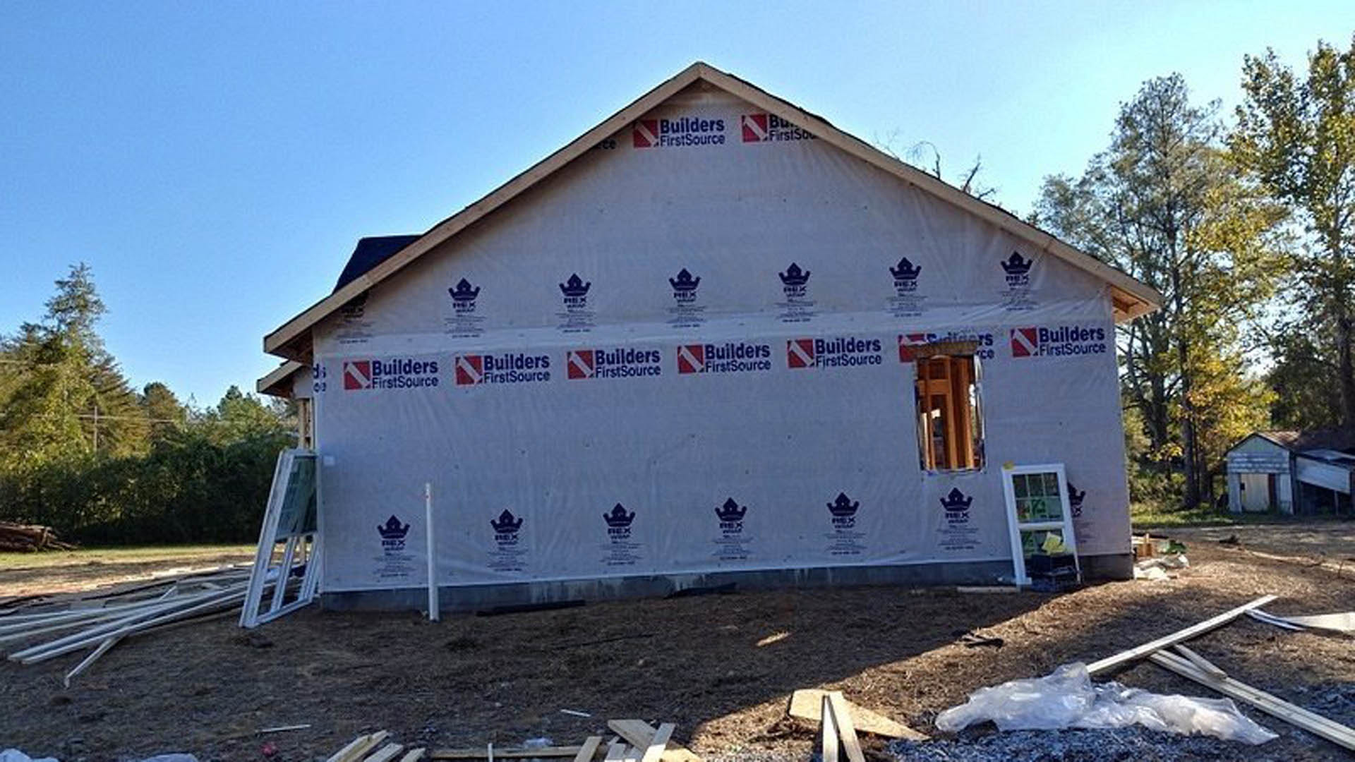 Partially built house wrapped in white tarp, exposed wooden framing, surrounded by dirt and trees under blue sky