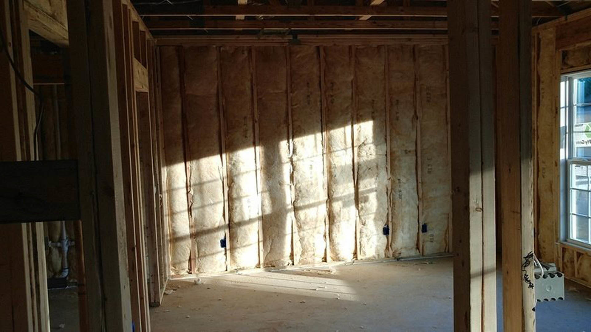 Room under construction with exposed wood framing, dense insulation filling wall cavities, concrete floor, window set in unfinished wall, and electrical box mounted near framing