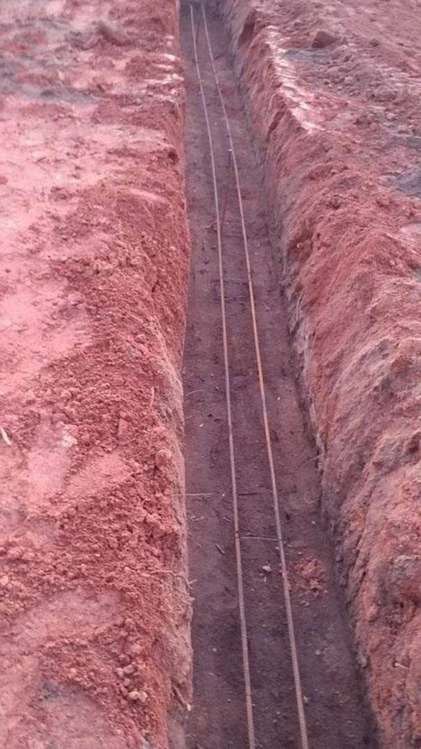 Outdoor trench dug in rocky ground with exposed electrical wires and metal rods, surrounded by mountain landscape
