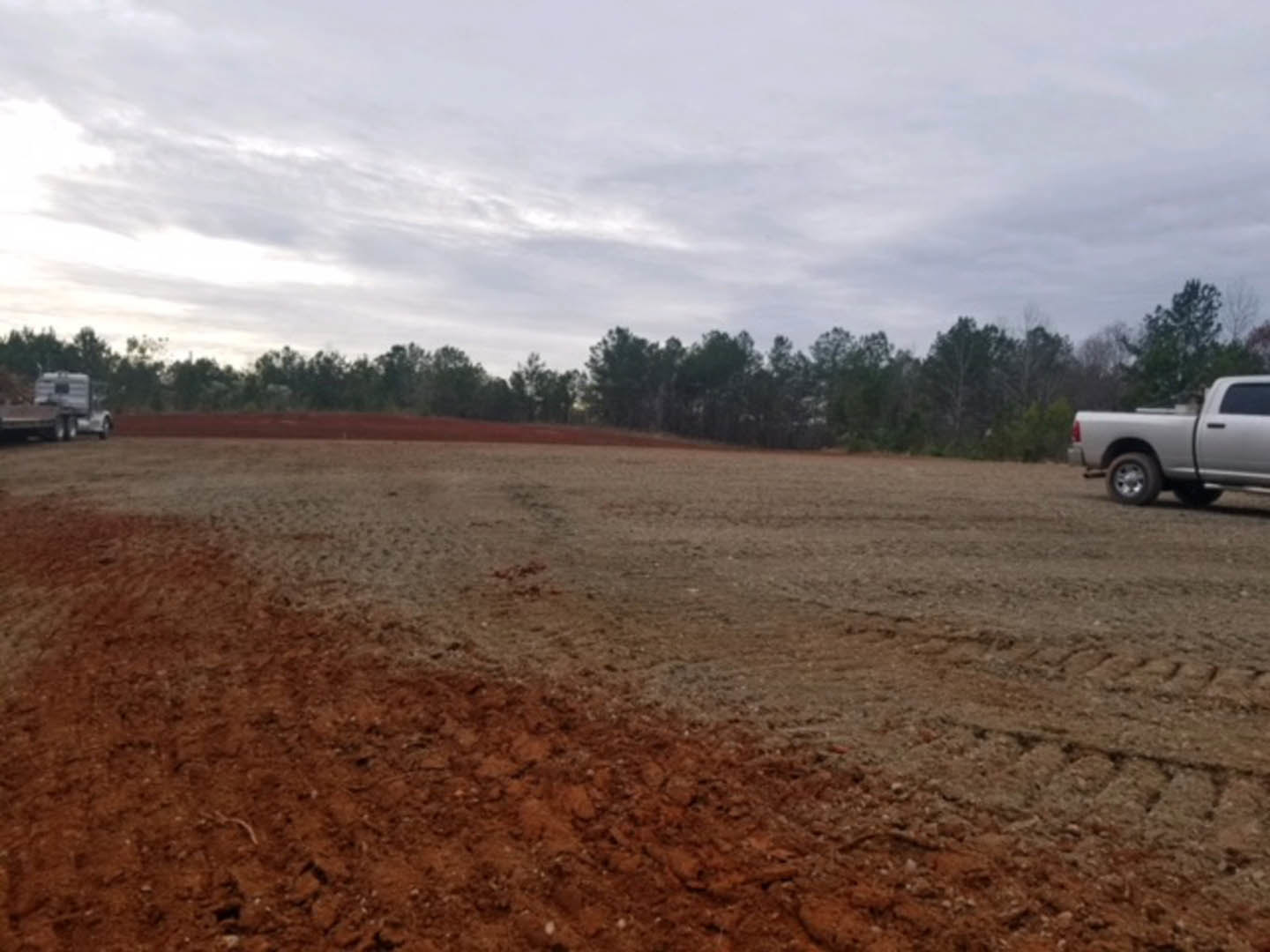 White truck with black door parked on dirt field, surrounded by trees under cloudy sky, close-up of tire visible.