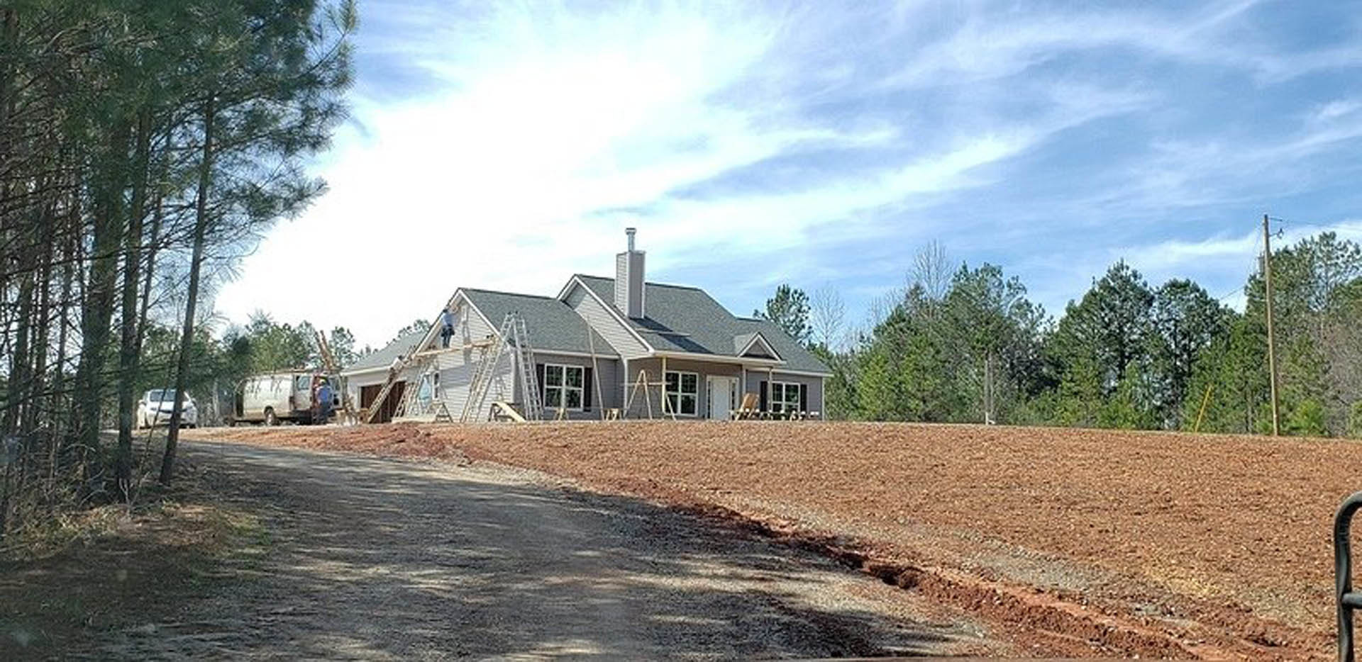 Partially built house with exposed framing, surrounded by dirt road and construction vehicles, set in rural landscape with trees and cloudy sky