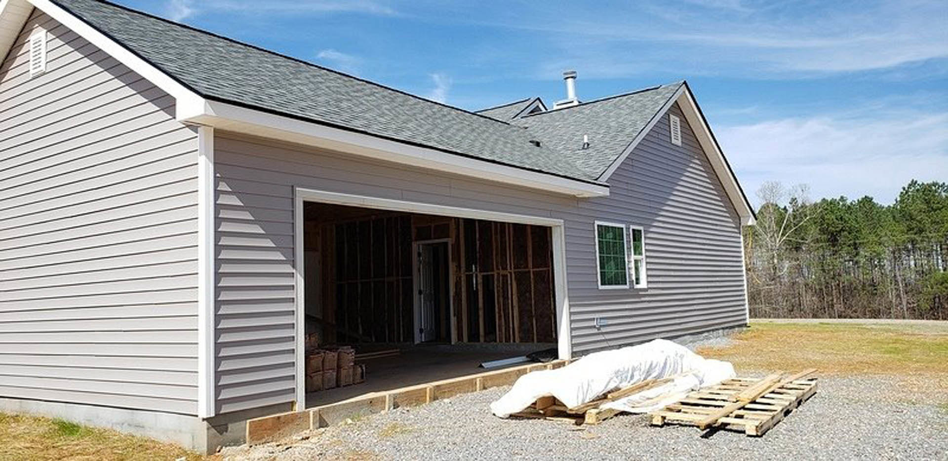 Framed house under construction with attached garage, exposed plywood siding, construction materials including pallets and barrels scattered in front, cloudy sky overhead