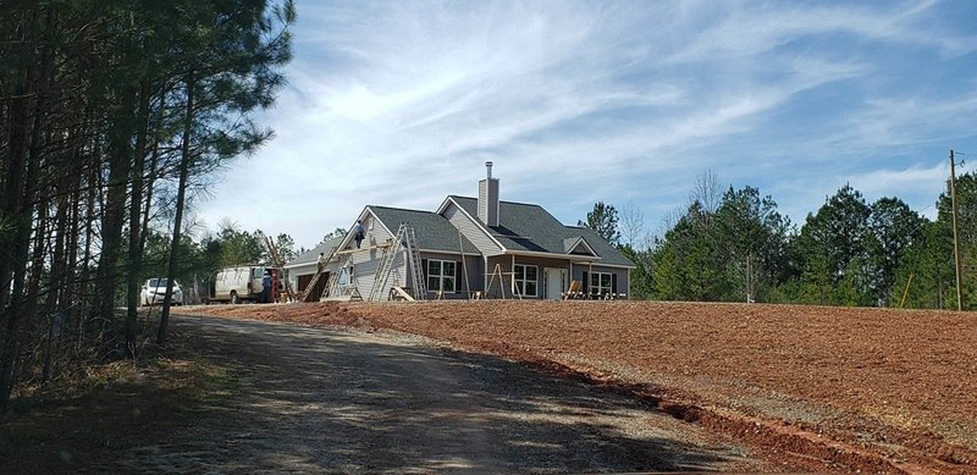 Wood-framed house under construction beside a paved road, surrounded by green trees; white van with open door and white car parked nearby