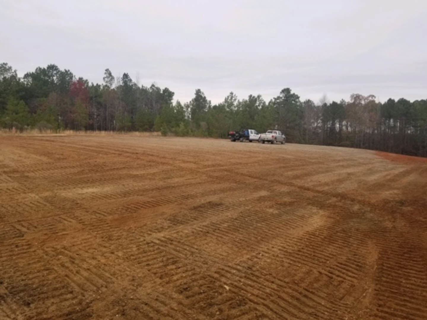 Expansive dirt field bordered by tall trees, two white trucks parked near the edge, open rural landscape under a clear sky