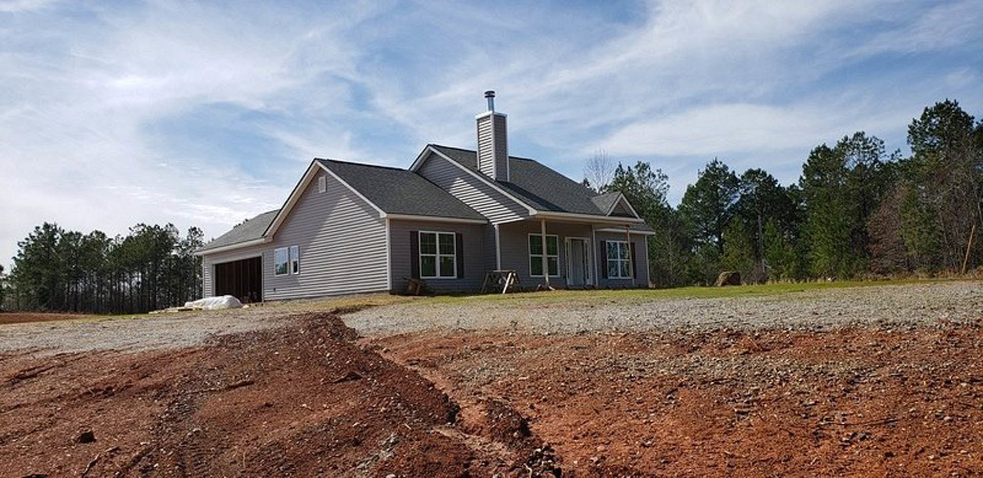 Two-story home with white-framed windows, gray siding, shingle roof, and a dirt patch in the front yard under a partly cloudy sky