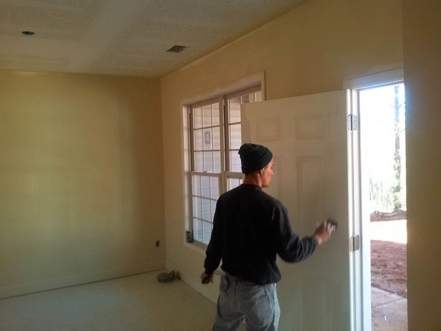 Man in black shirt painting white door near window with white frame in room with light-colored plaster walls and flooring