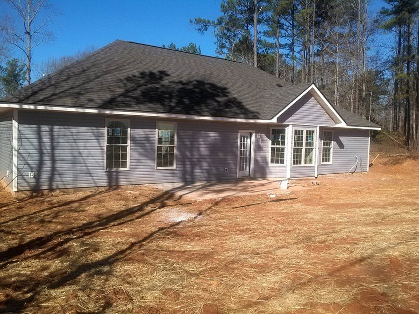 Two-story home with light siding, multi-pane windows, and a dirt yard bordered by a wooden fence; tree shadow falls across the ground, and a window displays a sign.