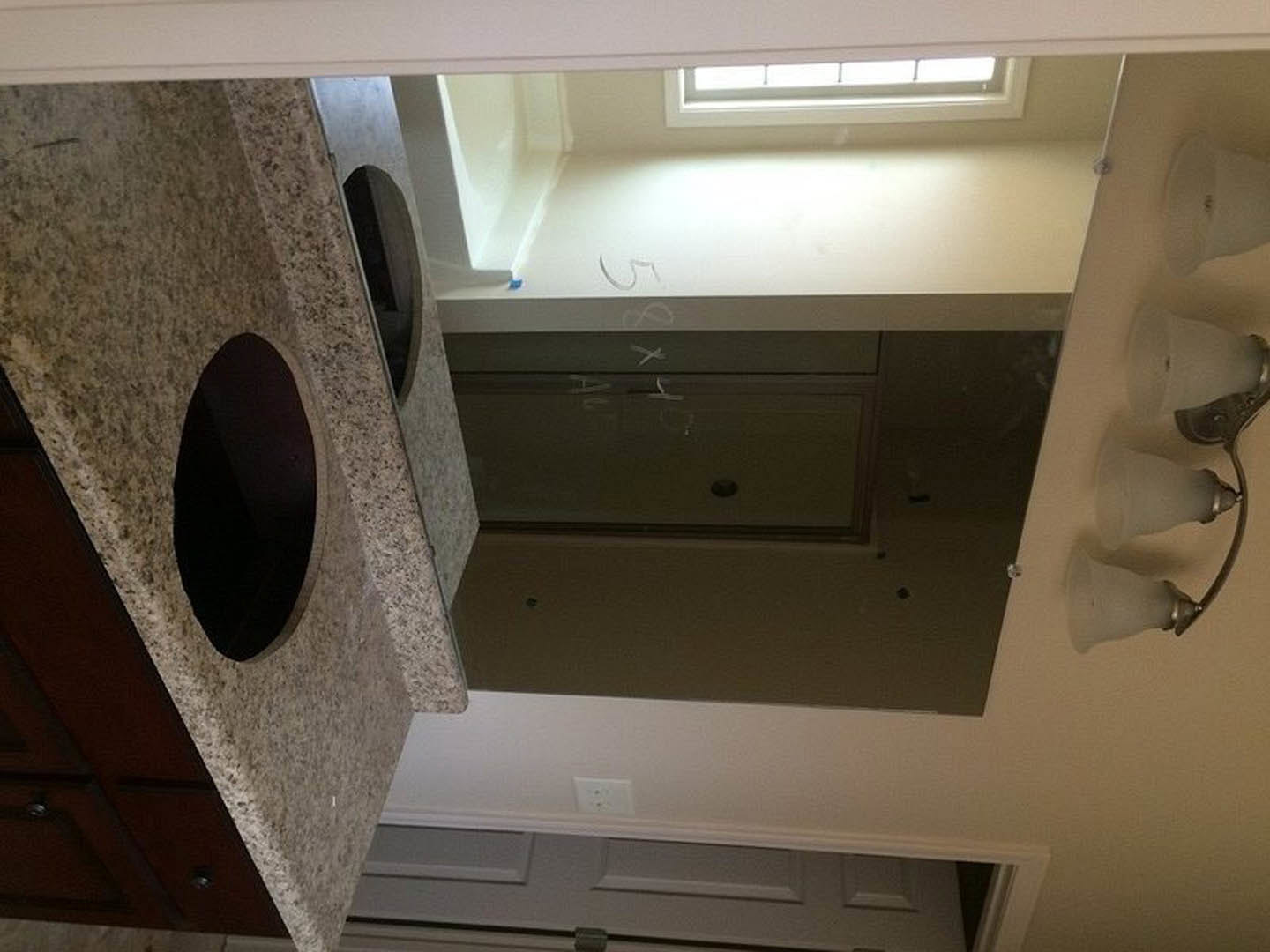 Modern kitchen featuring marble countertops, black circular sink, white plaster walls, large mirror above cabinetry, close-up of light fixture, window, and stone wall with circular