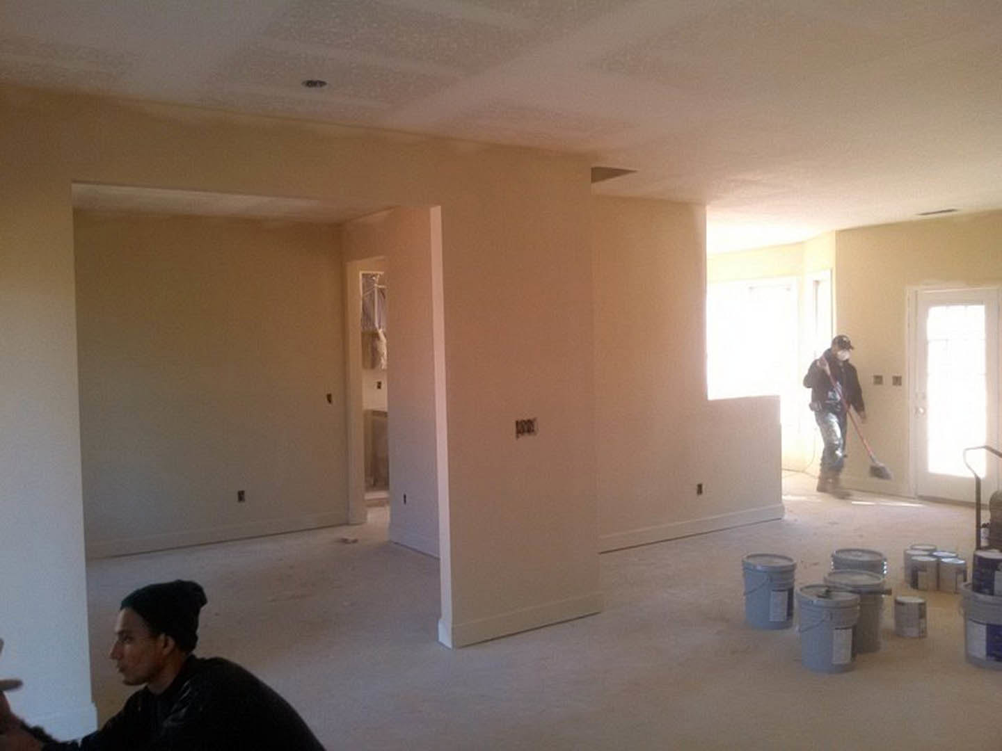 Two people painting a room with white walls, paint buckets and a broom on the floor, white door illuminated by bright light, trash can partially visible in the background.
