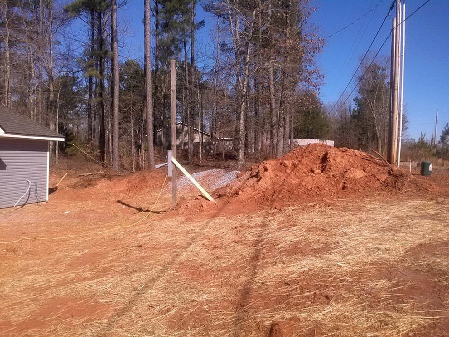 Large dirt pile in front of a partially visible house with gray roof, wooden post, and yellow rope; surrounding area includes soil, sparse plants, and open sky.