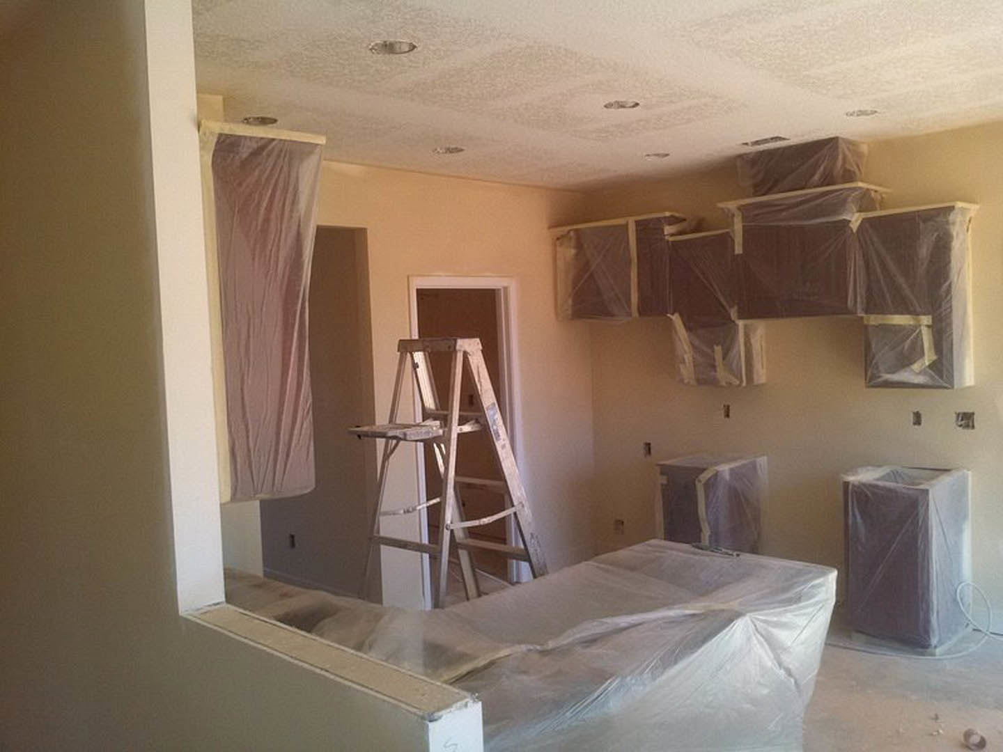 Bedroom under renovation with wooden ladder leaning against wall, bed covered in clear plastic, unfinished plaster walls, and construction materials scattered throughout.