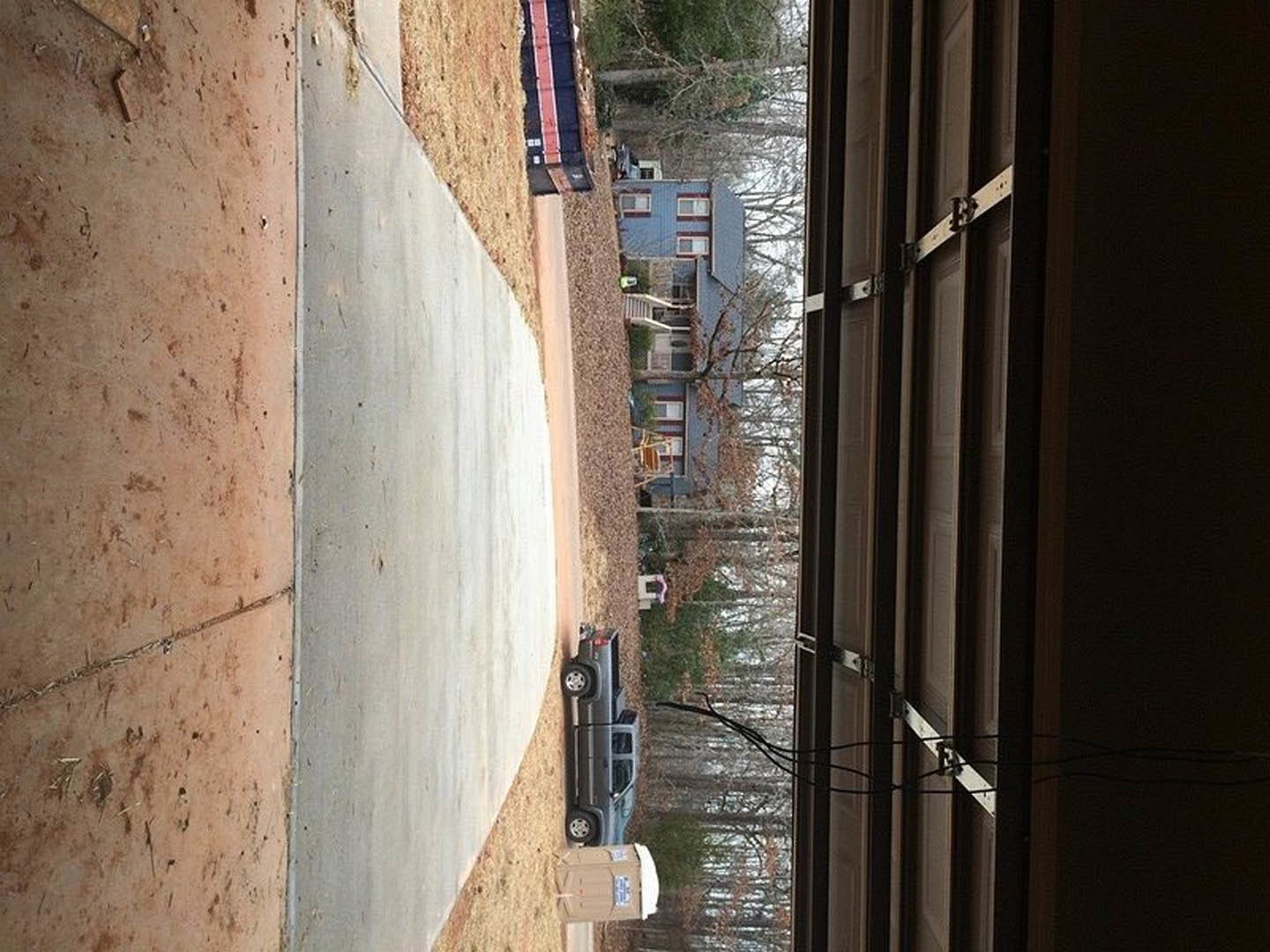White paneled garage door with a silver pickup truck parked on concrete driveway, leafless tree and brown mulch visible along side of home exterior