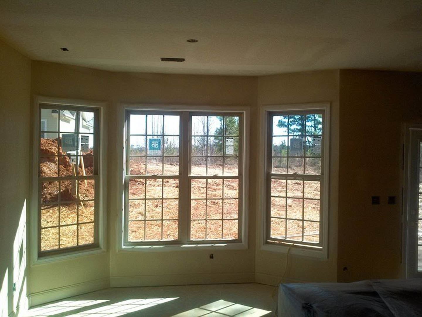 Bedroom with large windows, white walls, and a neatly made bed; blue and white sign visible on window, natural daylight illuminating space, outdoor view includes pile of dirt.