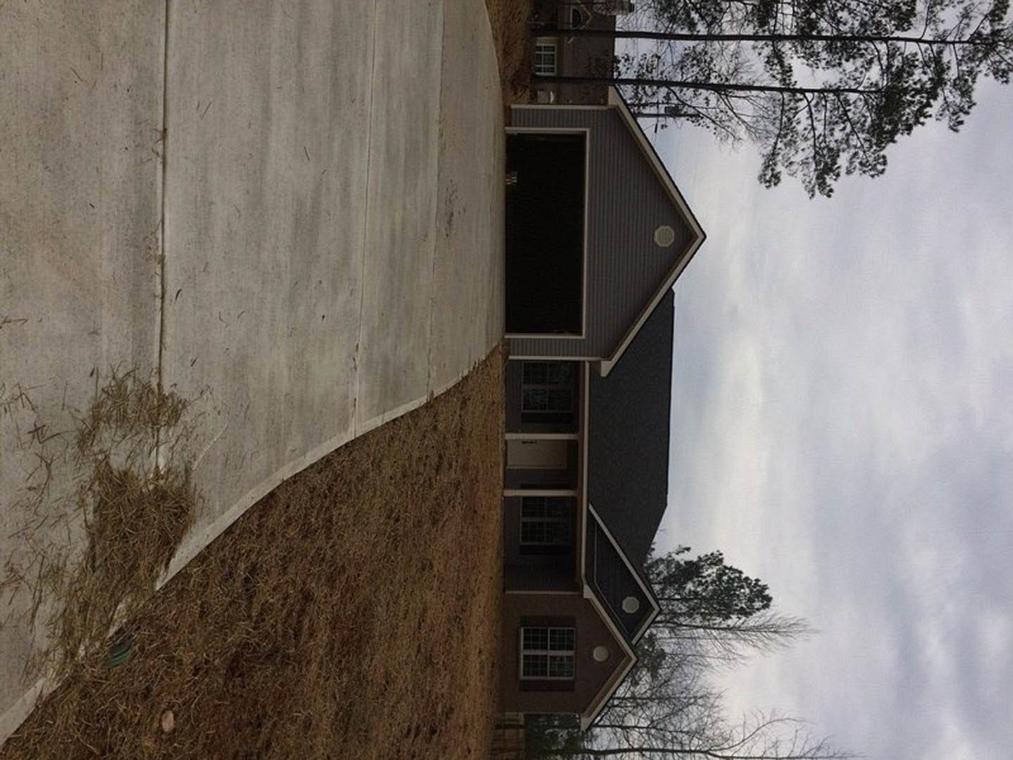 Two-story auburn brick home with black roof, open front door, large windows, concrete driveway, mature tree with spreading branches, partly cloudy sky