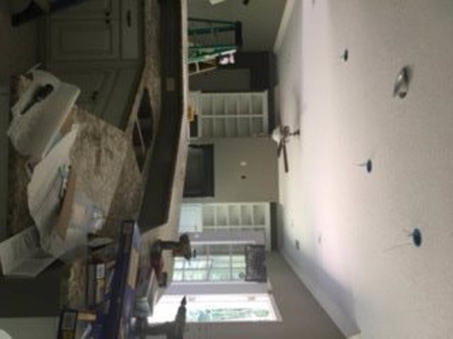 Modern kitchen with stainless steel sink, ceiling fan, white plaster walls, blue pins on one wall, light cabinetry, scattered papers on hardwood floor, and a close-up window with