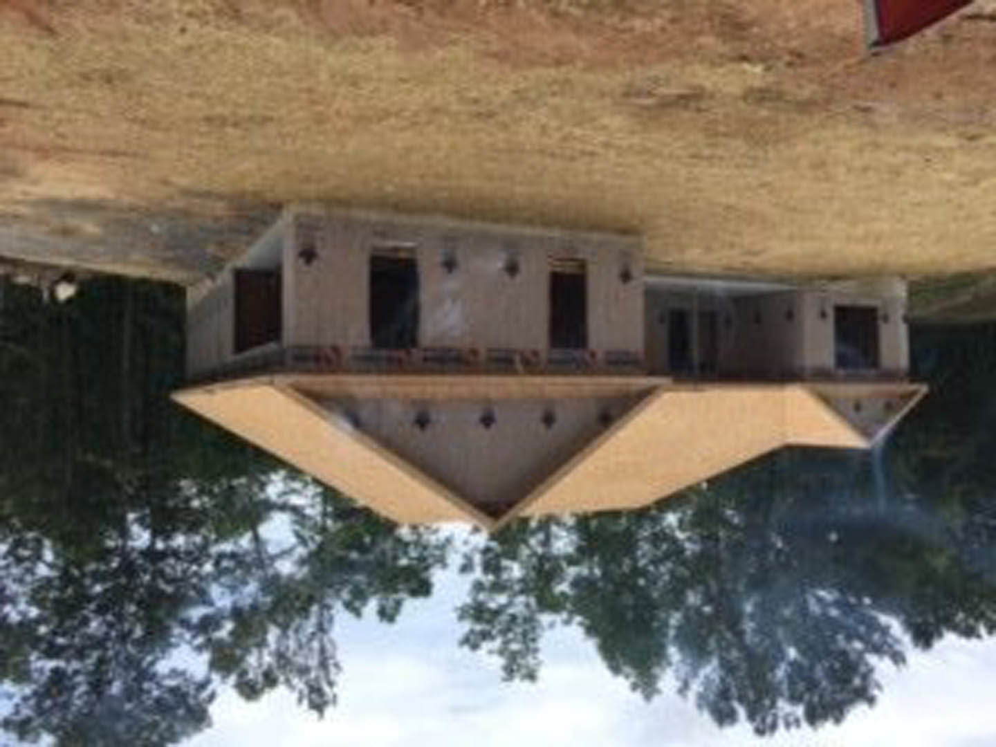 Two-story house with unfinished exterior, balcony, and large dirt patch in front yard, surrounded by sparse trees under a clear sky