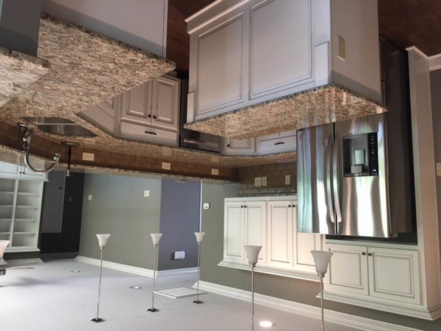 White kitchen cabinets and marble countertops, silver refrigerator with glass door, metal pole, modern lamp, box on counter, white wall, wood flooring, ceiling beams, large window.