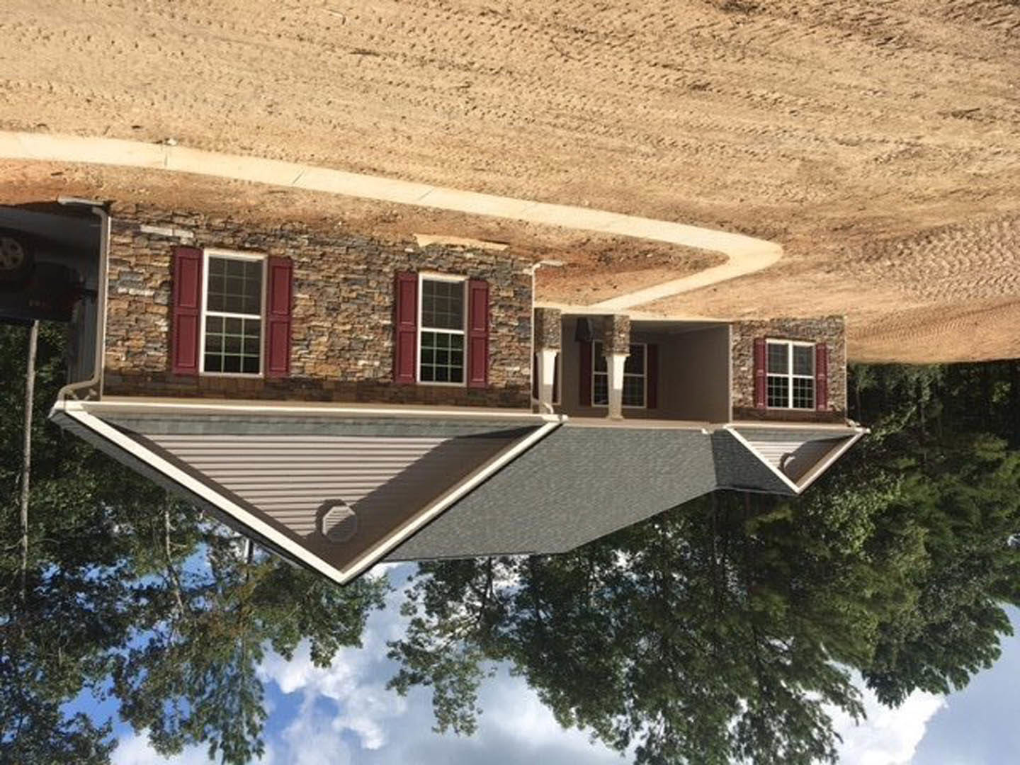 Two-story home with white-framed windows, brick and stone exterior walls, pitched roof, dirt driveway, and mature trees surrounding the property