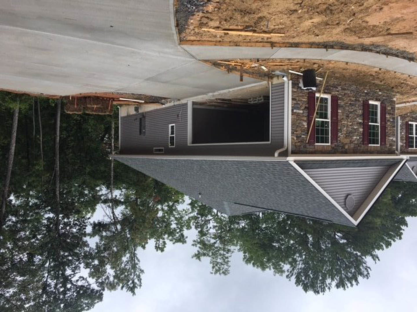 Partially built house with triangular roof vent, white-framed window, exposed dirt foundation, surrounded by trees and bordered by a road