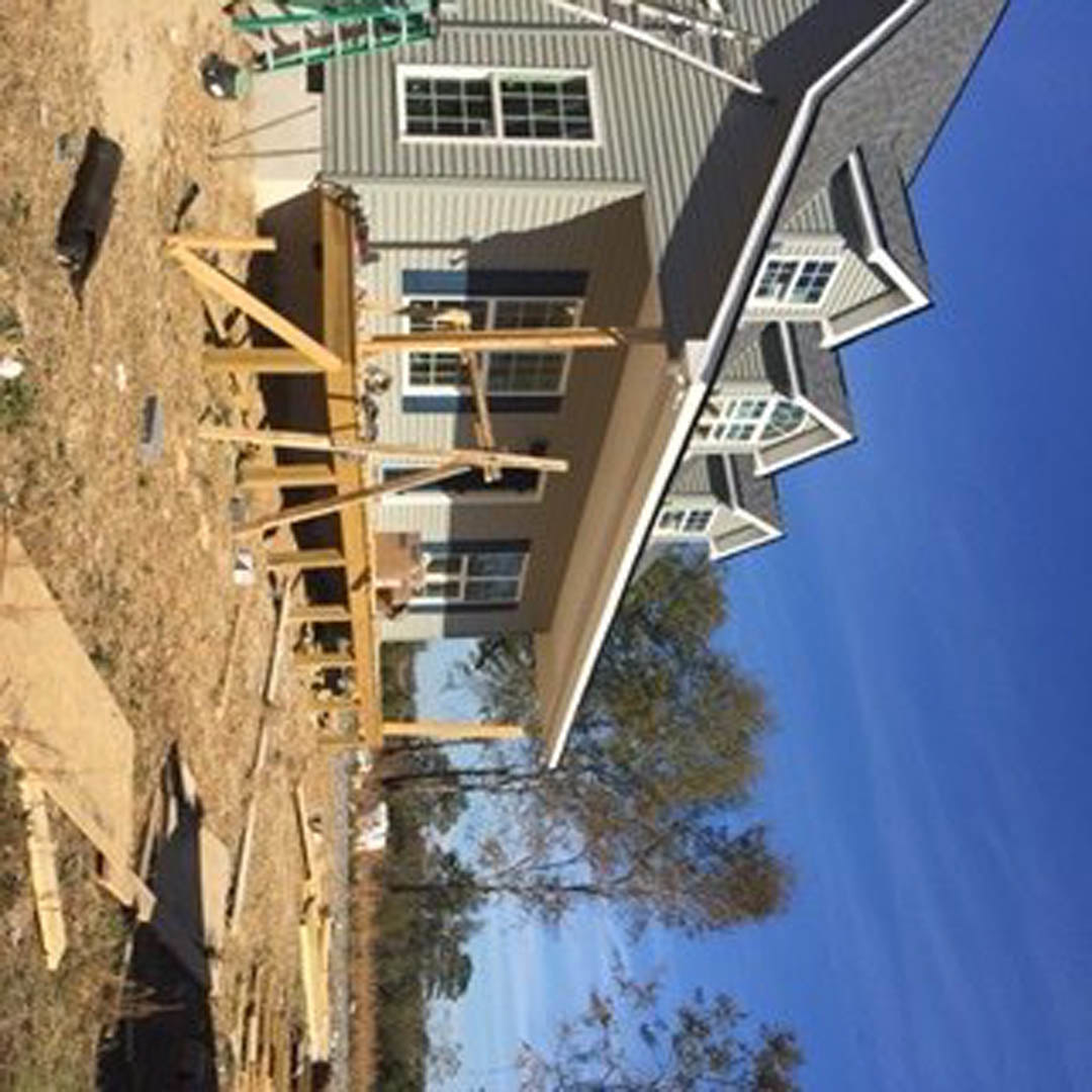 Partially built house with exposed framing, multi-pane window, pitched roof, tall tree nearby, metal ladder leaning against exterior wall, clear blue sky overhead