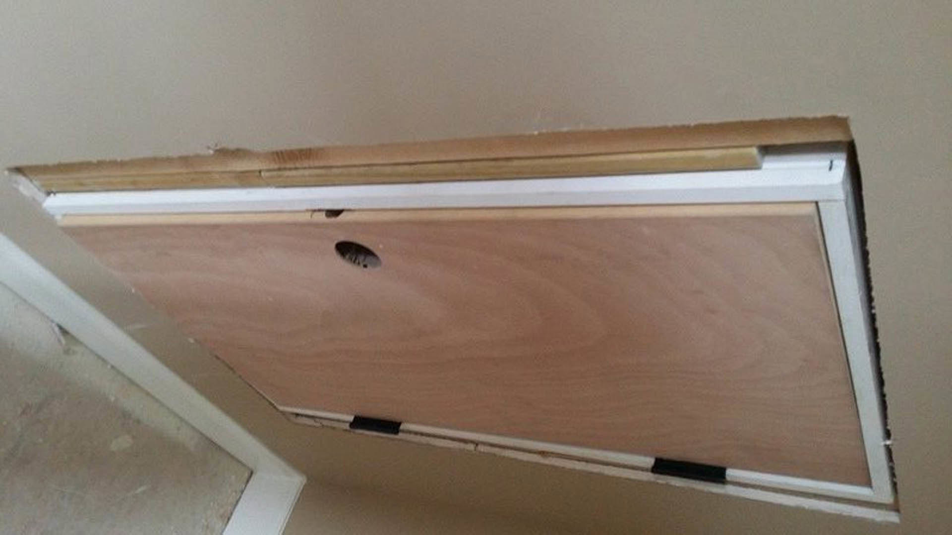 Wooden door mounted horizontally on a ceiling, surrounded by white walls and hardwood finishes in a modern interior room.