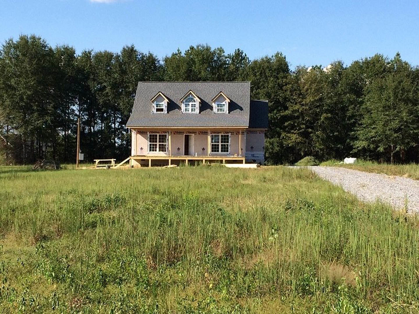 Two-story farmhouse with dormer windows, white siding, covered porch, gravel path bordered by tall grass, mature trees and cloudy sky in background