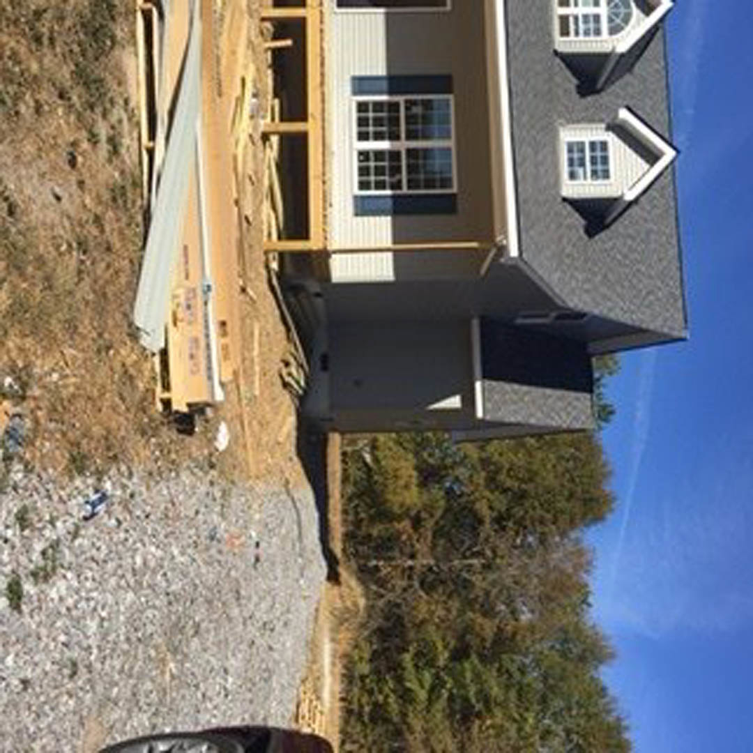 Partially built house with exposed framing, white-trimmed windows, star decoration on glass, dirt road in foreground, parked car, leafy tree nearby, clear sky overhead