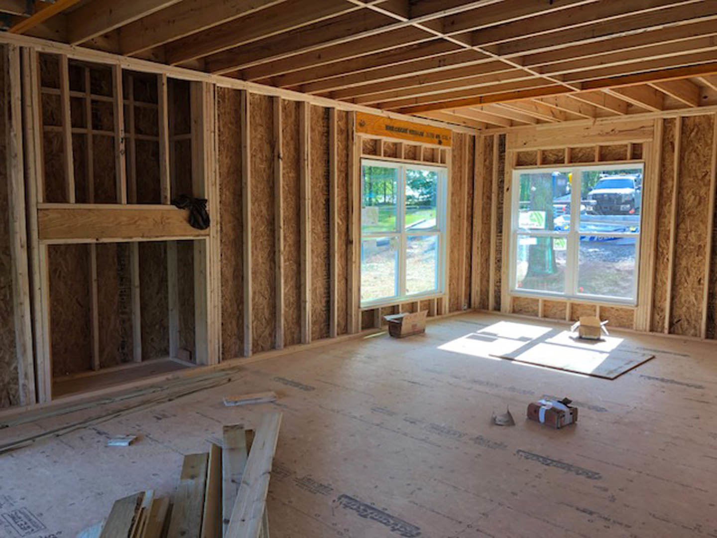 Living room with exposed wood ceiling beams, large windows, unfinished wood planks stacked on the floor, and a window overlooking grassy yard