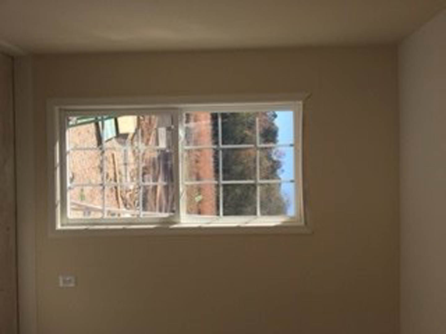 Large window with white trim set in a plaster wall, sunlight filtering through blinds, leafy trees visible outside