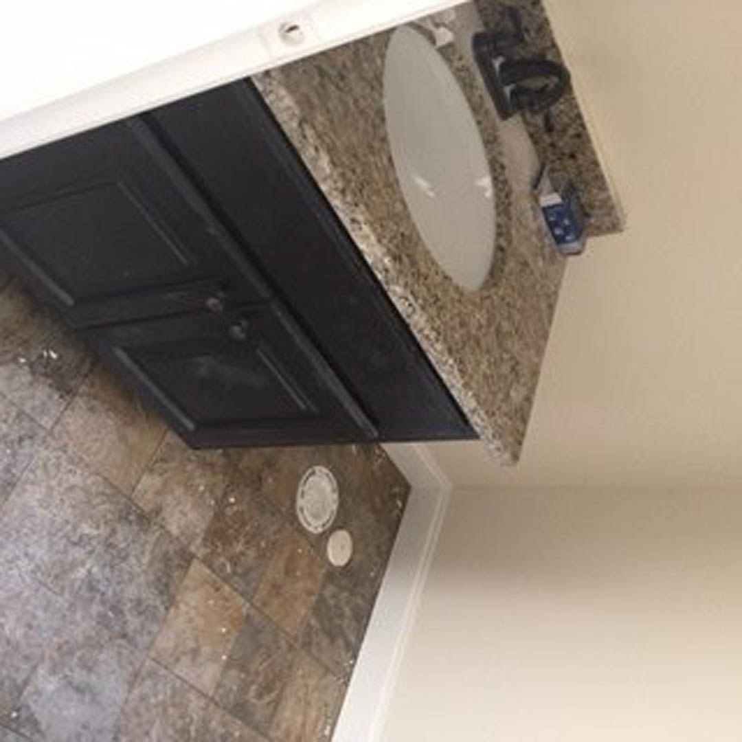 White ceramic sink set in a light wood vanity cabinet, rectangular wall mirror above, pale tile floor, white plaster walls, chrome faucet, partial view of toilet.