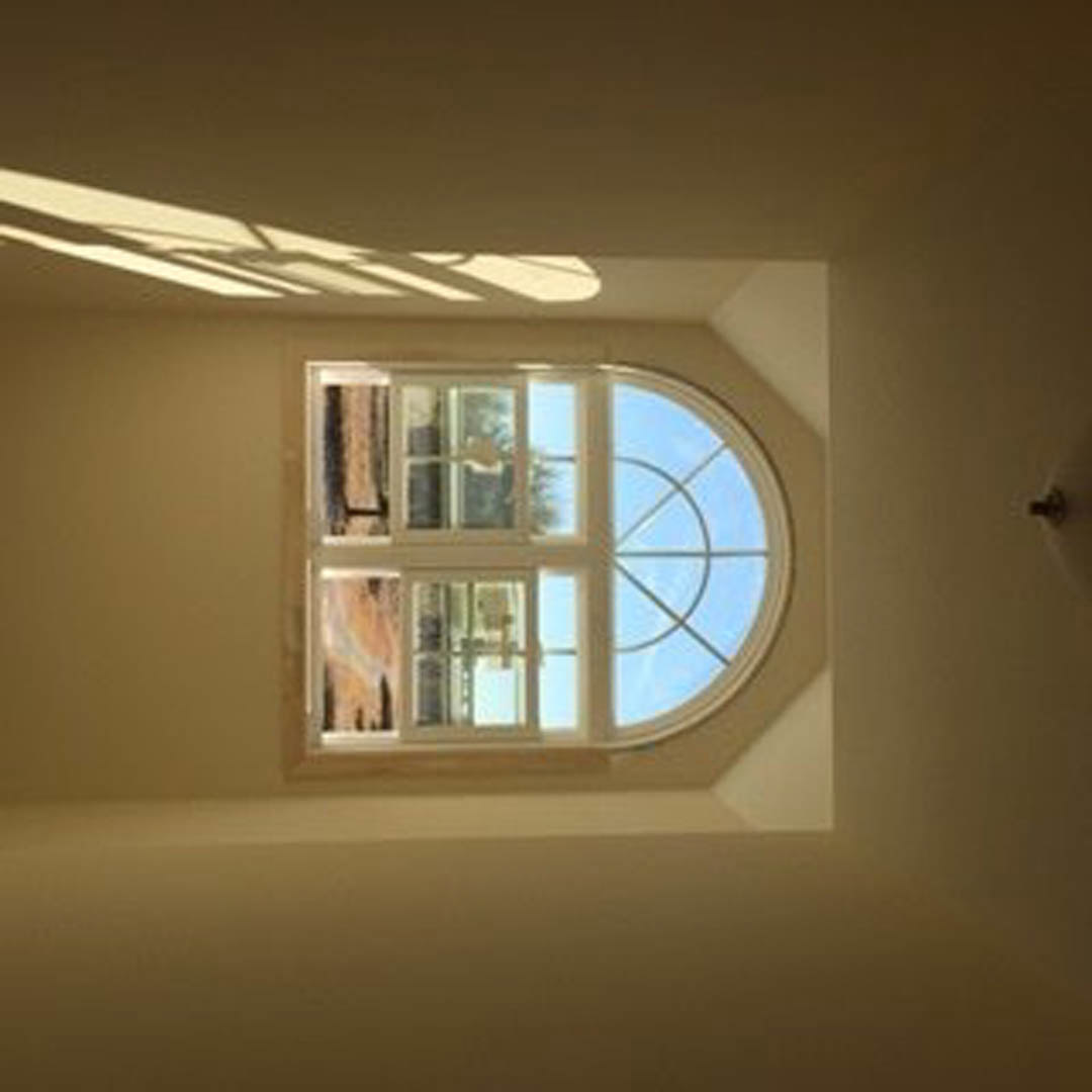 Large rectangular window with circular transom, white plaster walls, sunlight casting geometric shadows across interior room
