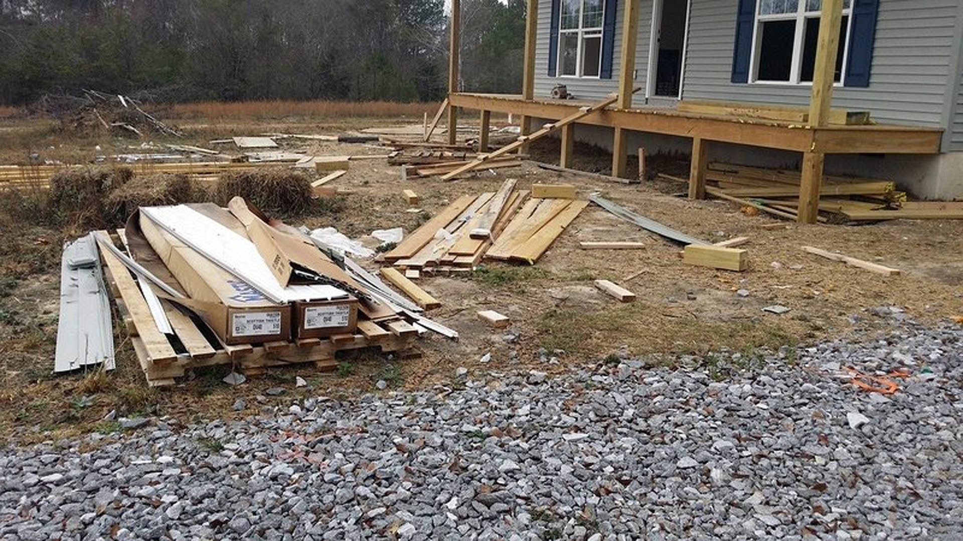 Partially constructed wooden house with exposed framing, unfinished wooden deck, piles of lumber and rocks on the ground, and cardboard boxes stacked on pallets