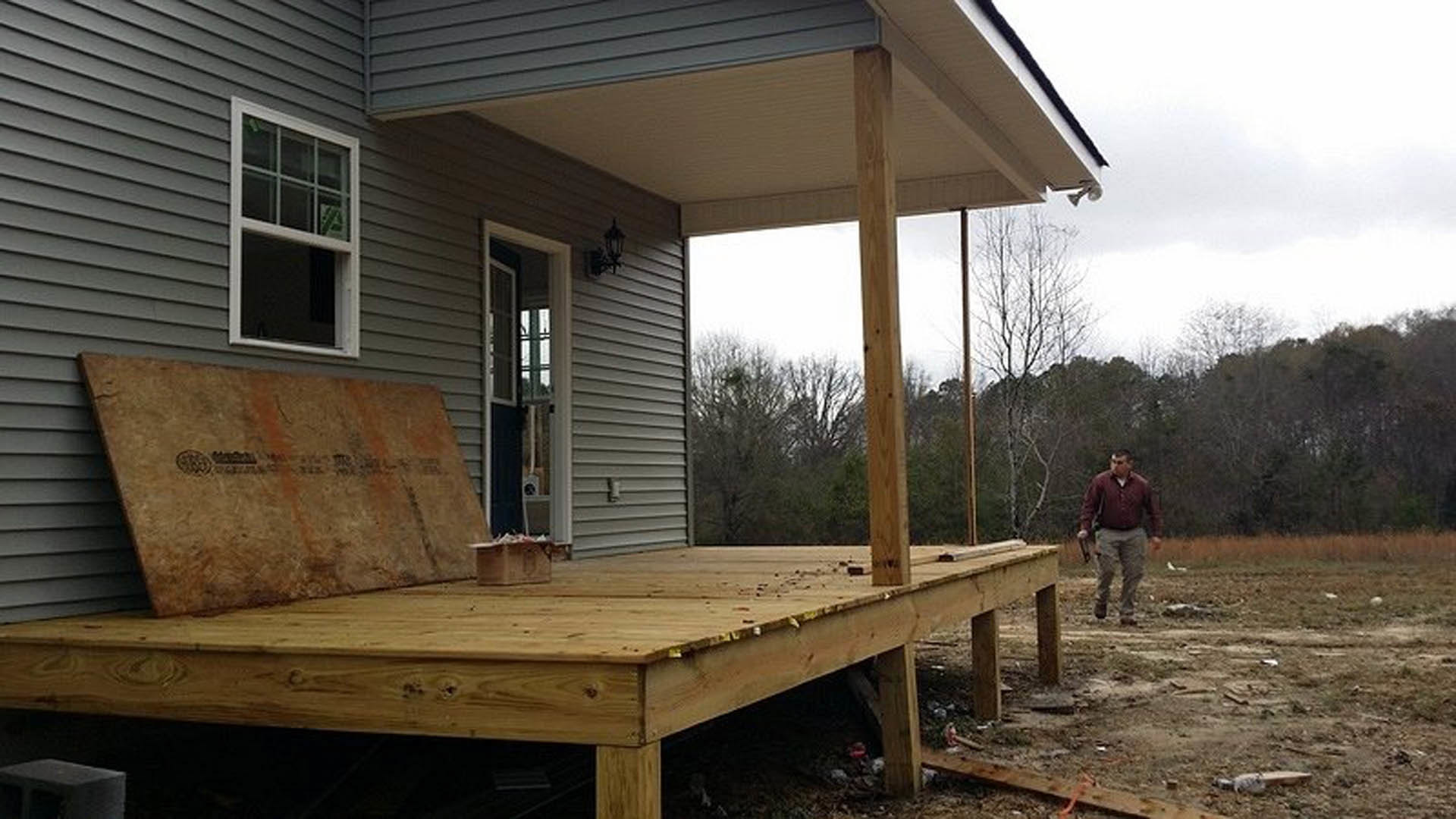 Man in red shirt standing on wooden deck beside house, large window visible, surrounded by trees and shaded outdoor area