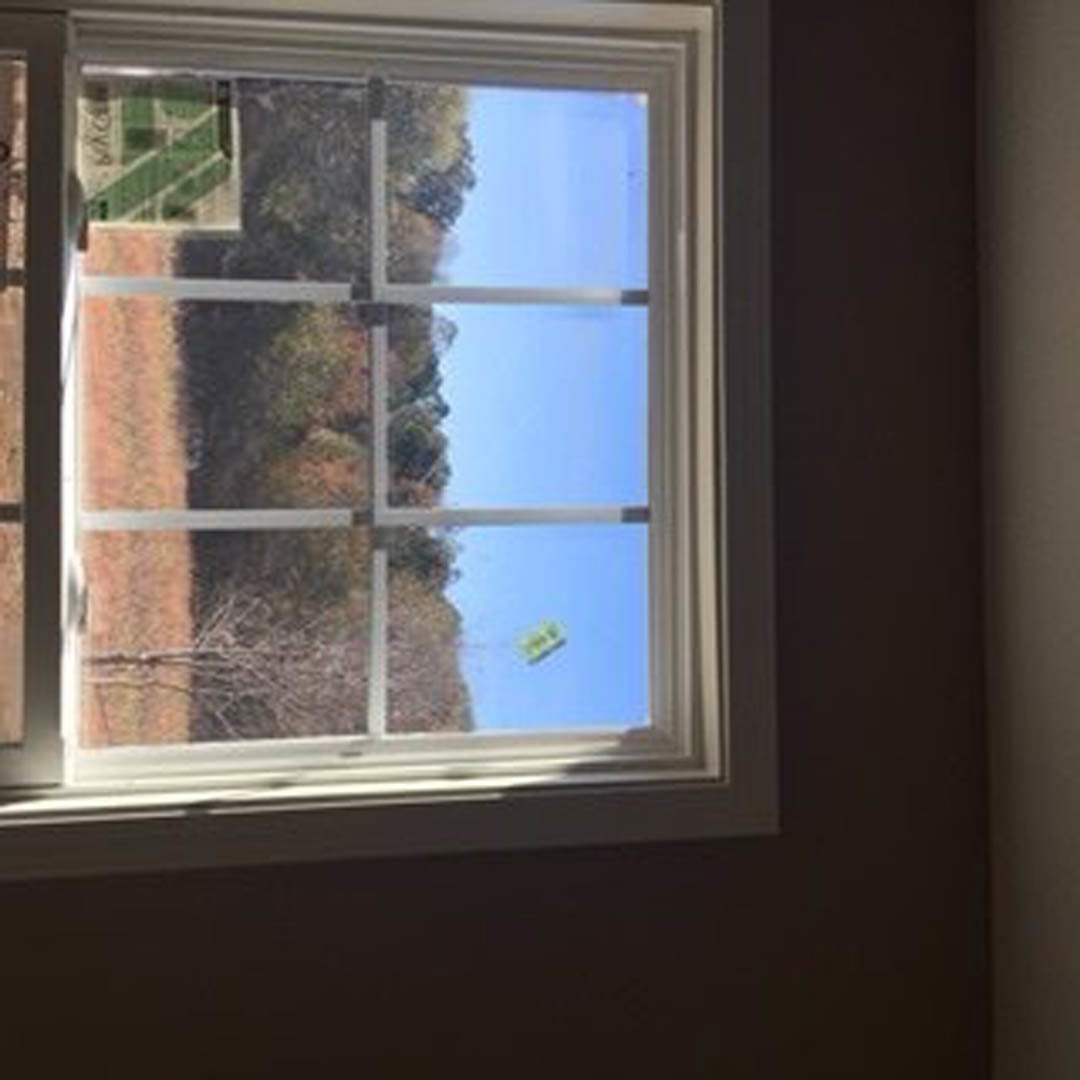 Large window framed by white trim overlooking a grassy field, tennis court, and scattered trees, with sunlight illuminating the indoor wall.