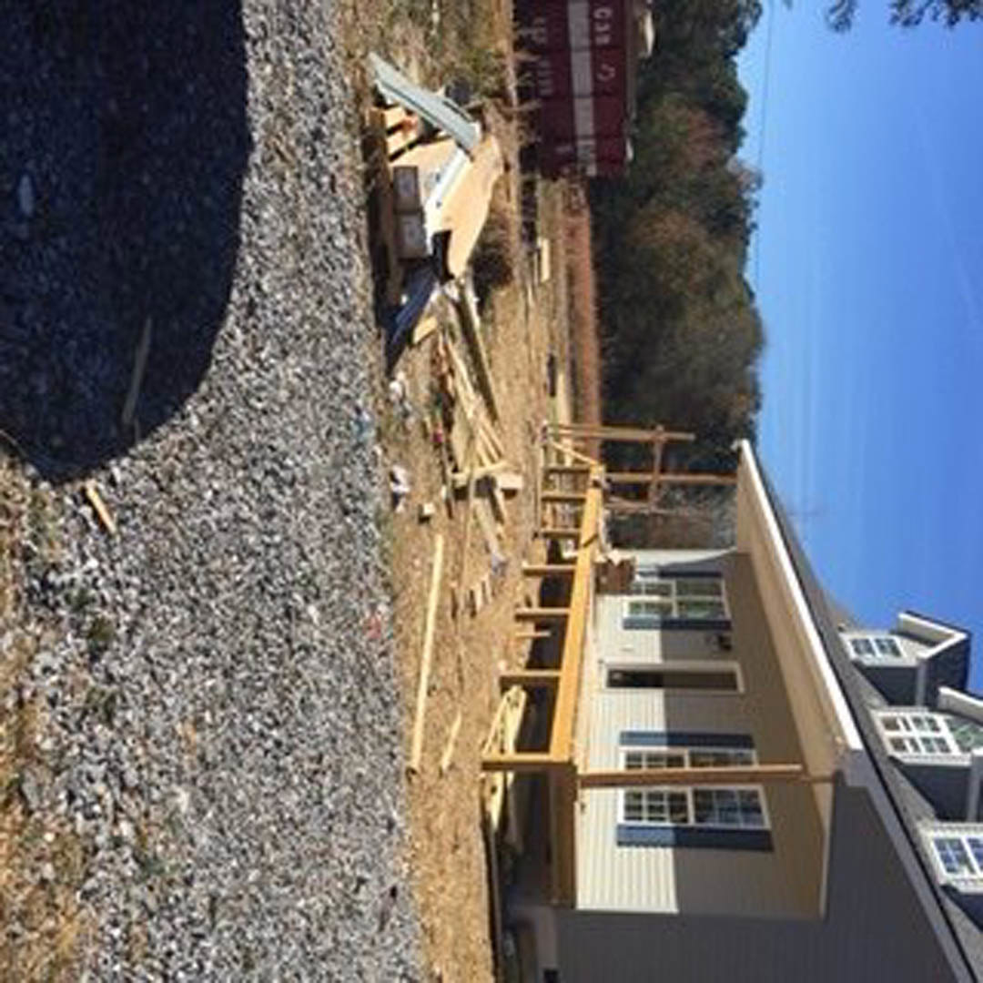 Two-story house under construction with exposed wooden framing, roof partially finished, gravel driveway, red and white construction sign, ladder leaning against exterior wall