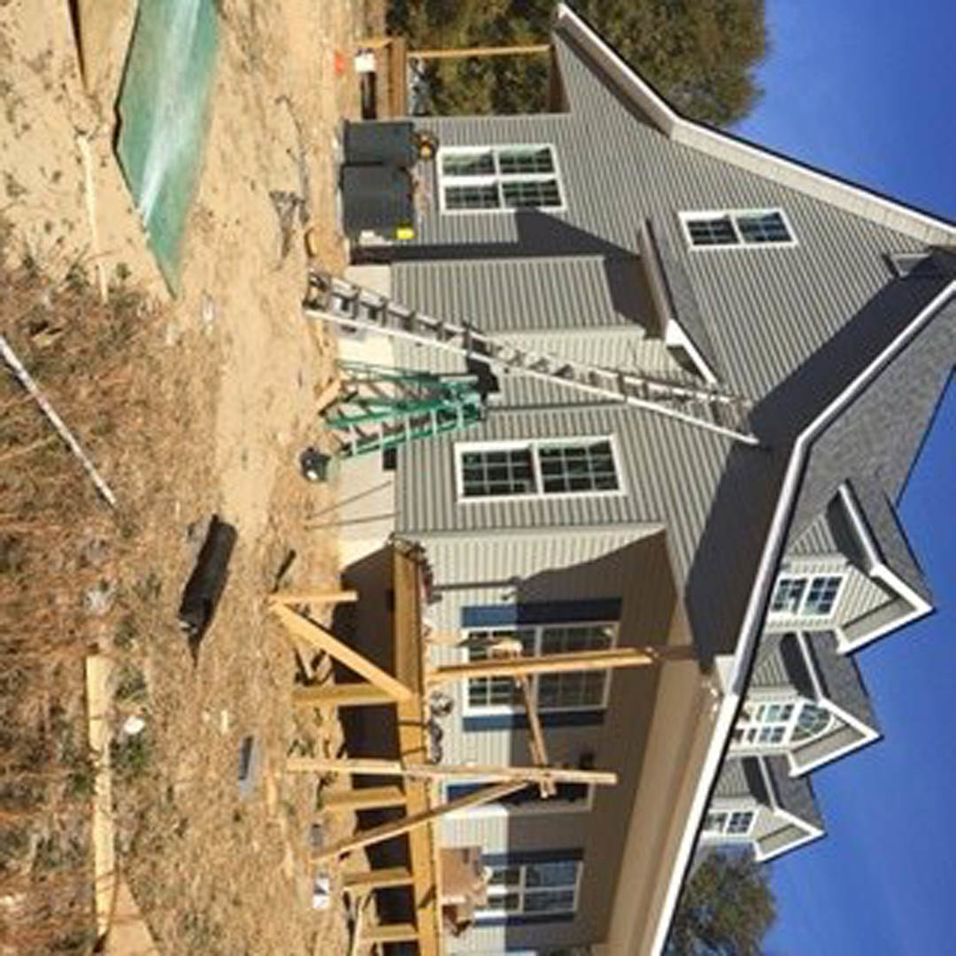 Partially built house with exposed wooden framing, white window frames, ladders leaning against exterior, construction materials scattered on ground, tree visible in background