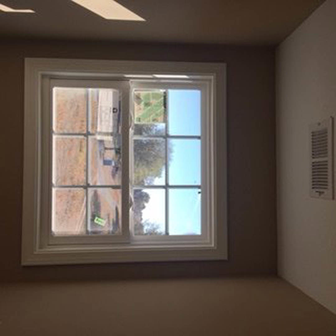 Large window with white trim set in plaster wall, sunlight filtering through sheer blinds, vent installed below windowsill, view of grassy field outside
