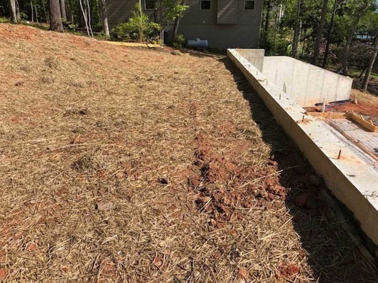 Concrete foundation beside a house with dirt ground and trees in the background