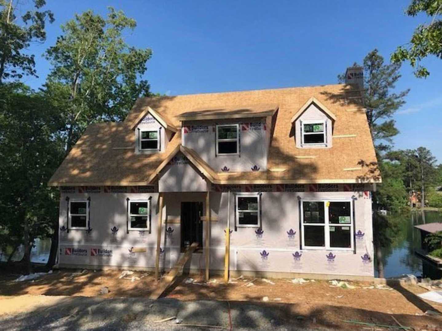 Framed house under construction with exposed wood, white window trim, porch area, and leafy trees in the background under a blue sky