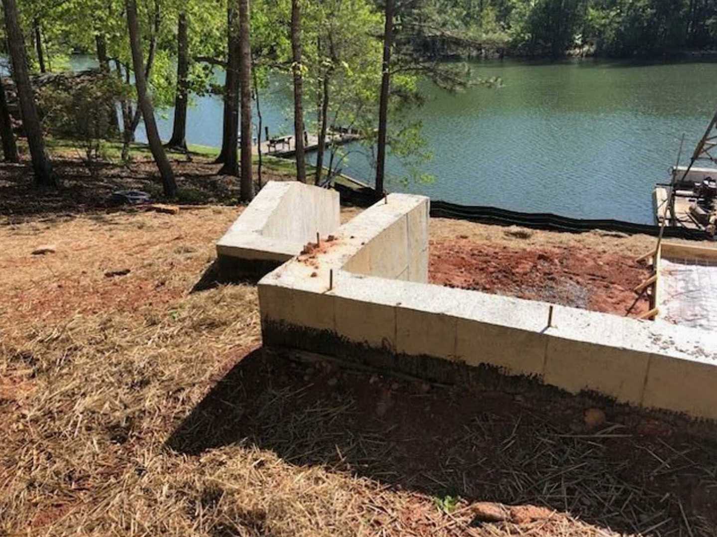 Concrete block wall beside a lake, surrounded by grass, trees, and plants, with a metal fence and wooden bench nearby.